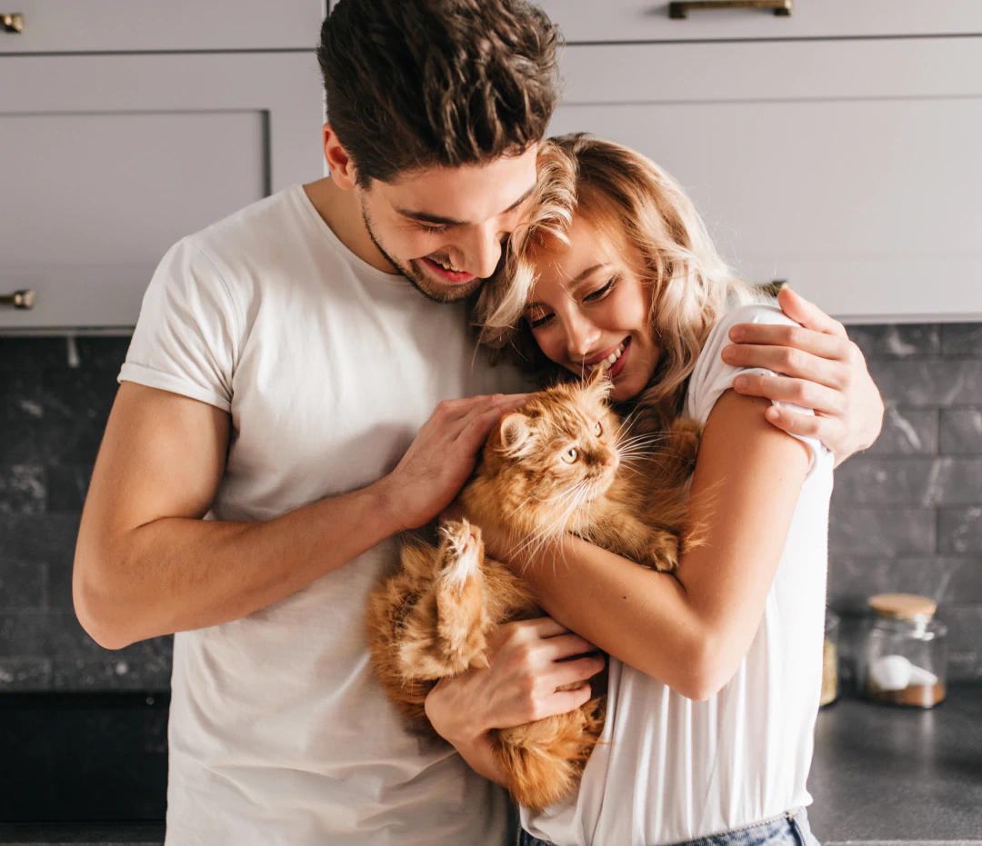 Desktop_Contact_Us_Always_Here A smiling couple in a kitchen hugs each other while holding a fluffy orange cat, which the woman is petting.