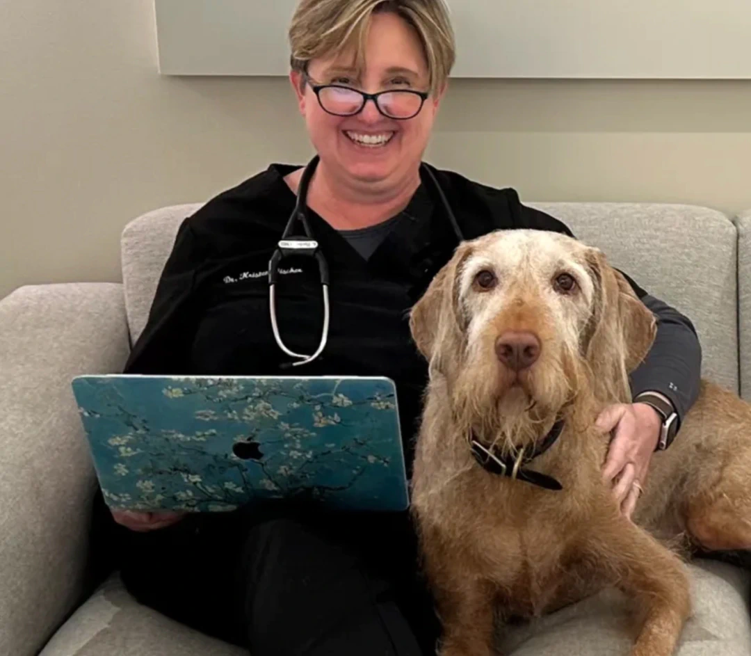 Desktop_Meet_the_Doctor_Meet_Kristen A smiling person in scrubs with a stethoscope, holding a laptop, sits on a couch next to a large, brown dog.
